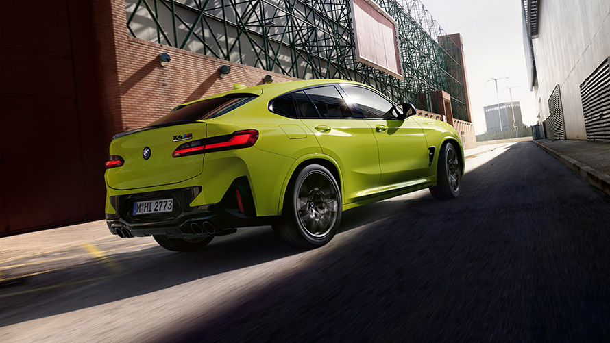 BMW X4 M Competition, F98, LCI, atjaunināts, 2021, Sao Paulo Yellow krāsa, trīs ceturtdaļu priekšpuses skats, braucienā starp rūpnīcas ēkām