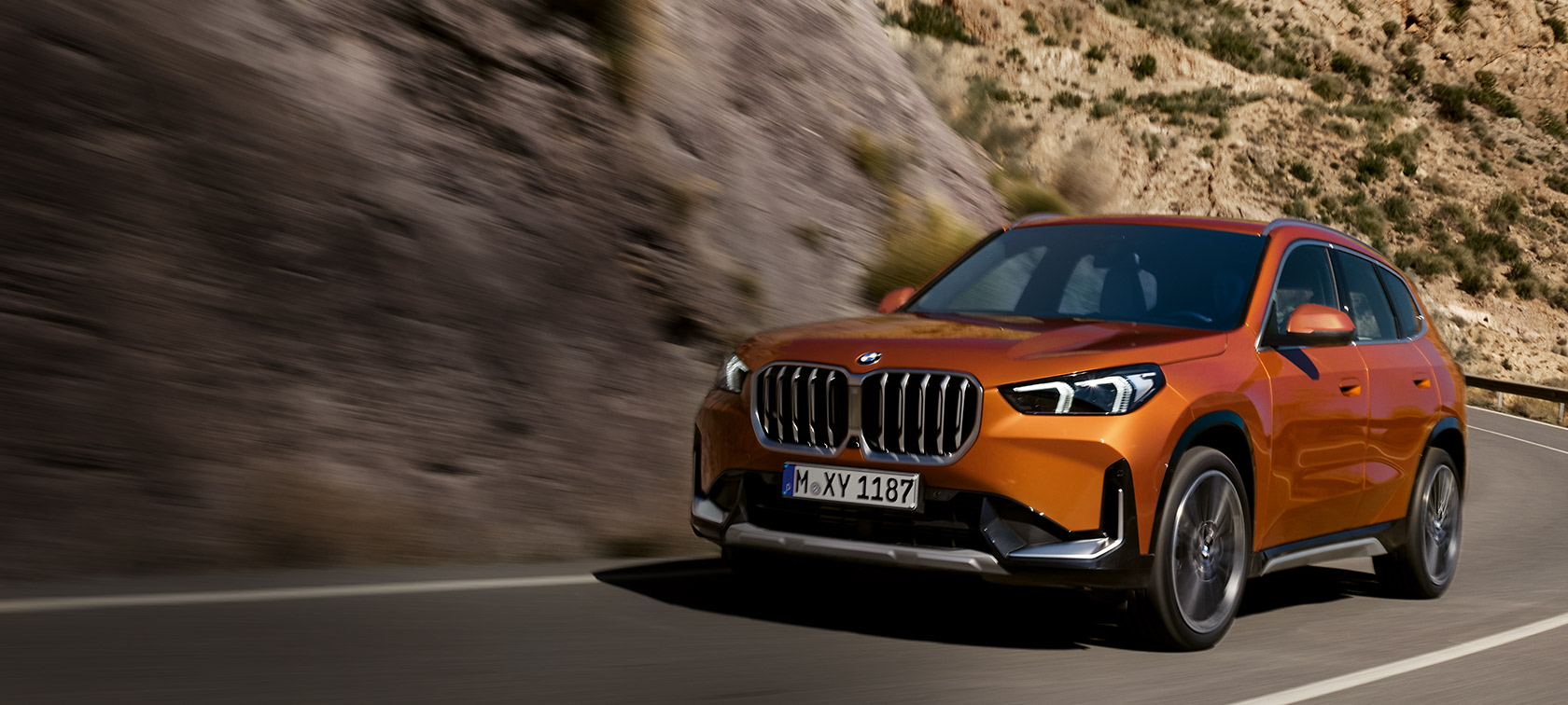 BMW X1 U11, xLine, metāliska Utah Orange krāsa, trīs ceturtdaļu priekšpuses skats, braucienā pa lauku ceļu